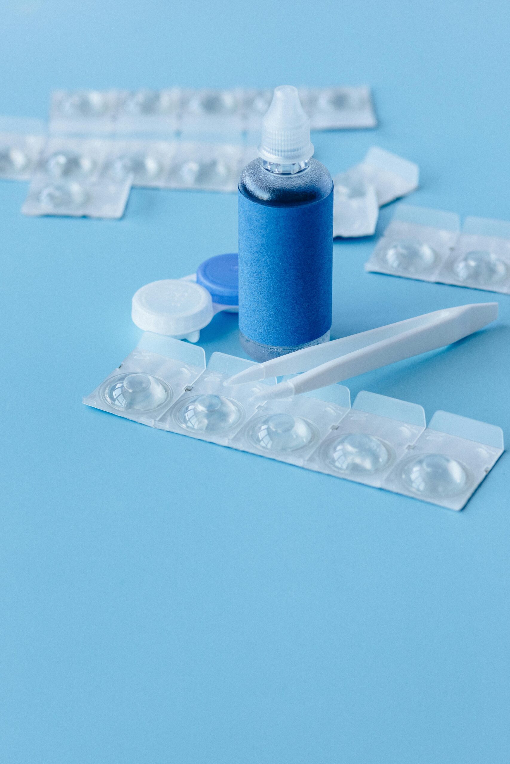 A close-up of contact lens care items including solution, blister packs, and tweezers on a blue background.